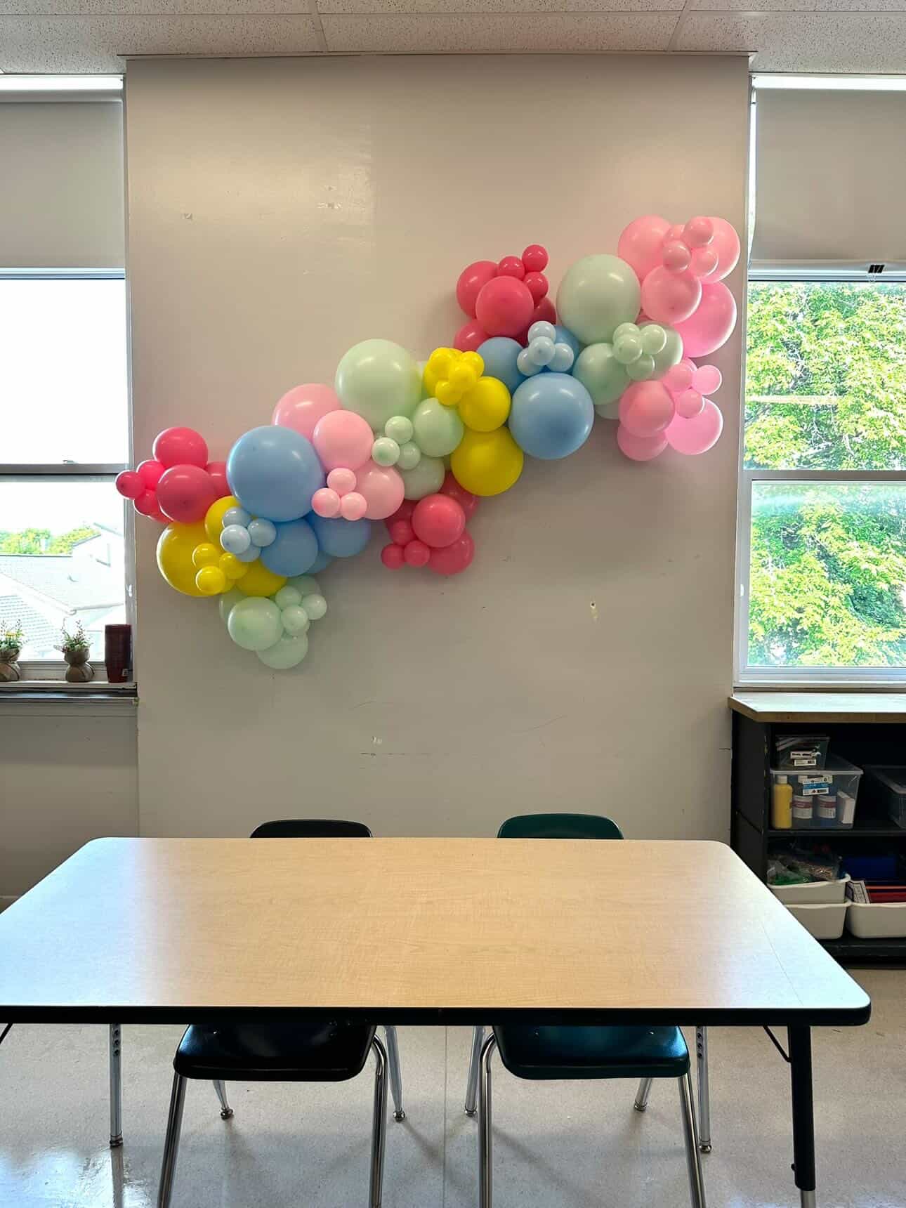 Pastel rainbow balloon garland hung on a classroom wall above a table, Winnipeg